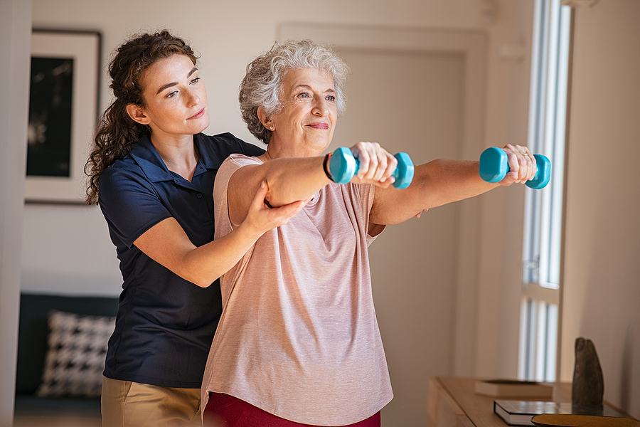 Old woman training with physiotherapist using dumbbells at home. Therapist assisting senior woman with exercises in nursing home. Elderly patient using dumbbells with outstretched arms.